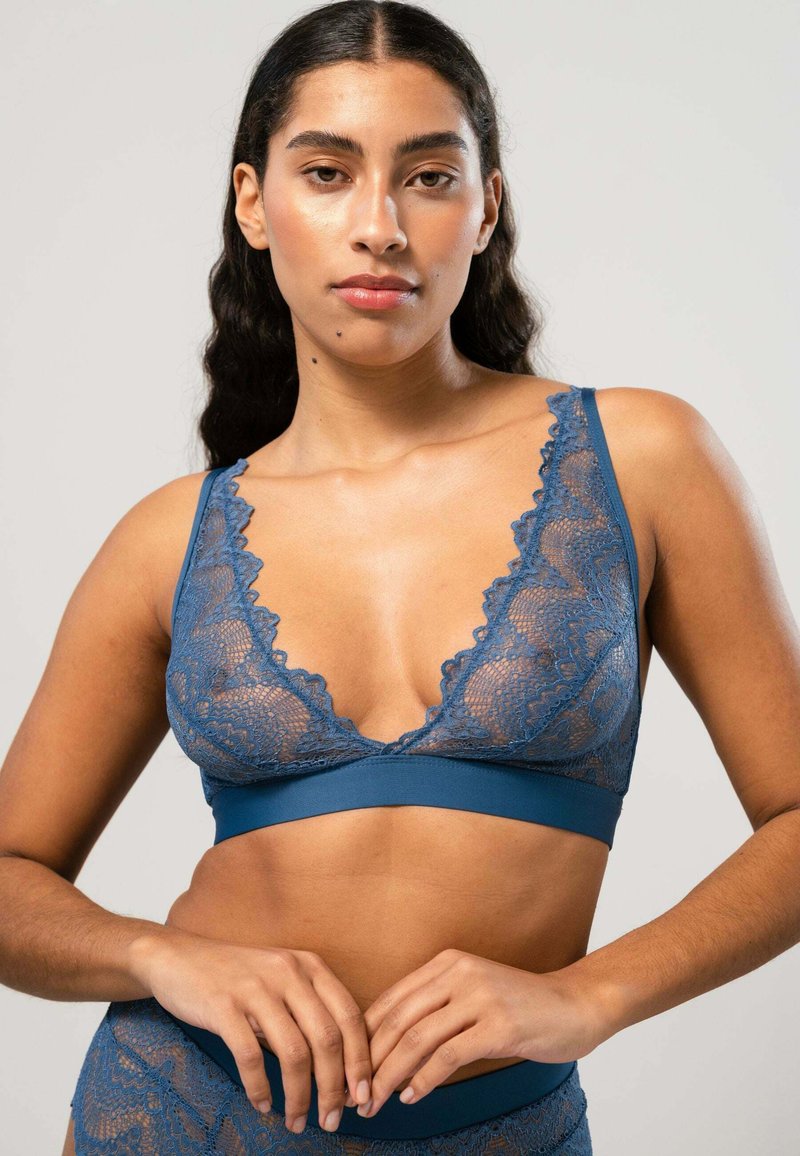 Spets-bh i djupblått, med en djup urringning med vågiga kanter och ett fast band nedanför. Modellen poserar med händerna sammanflätade.