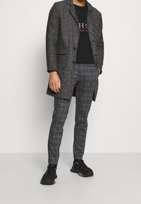 Abrigo gris de espiga sobre una camiseta negra, combinado con pantalones de cuadros grises y zapatillas negras. Las texturas notables incluyen los patrones de espiga y cuadros.