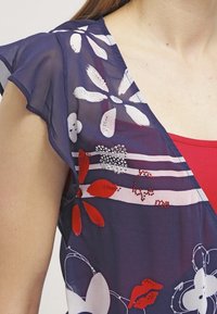 Genomskinlig marinblå blus med blommönster i röd och vit, kapärmar och dekorationer. Har en v-ringning och en lätt textur.