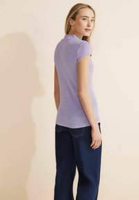 Kortärmad lila polo med en strukturerad, glittrande textil; figurnära vid midjan, tillsammans med mörkblå jeans, sedd bakifrån.