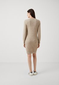 Abito beige a maglia con maniche lunghe e scollatura rotonda, silhouette aderente e orlo a costine, indossato con sneakers bianche con dettagli neri.