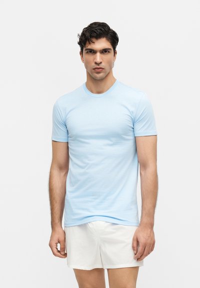 Άνδρας όρθιος, φοράει ανοιχτό μπλε κοντομάνικο μπλουζάκι και λευκά σορτς, σε απλό λευκό φόντο.