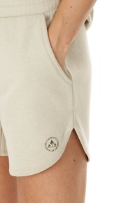 Ljusbeiga bomullsshorts med sidfickor, med en snedställd fåll och ett logotypmärke i botten, mjuk textur och avslappnad design.