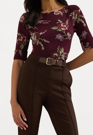Kvinde iført en vinrød blomstret top med albuelange ærmer, kombineret med højtaljede mørkebrune bukser og et matchende bælte.