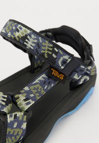 Teva sandalias musta tallaga, sinise aktsentide ja reguleeritavate mustritega rihmadega tumesinises, rohelises ja beežis. Rihmal on märkimisväärne logo.