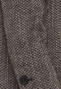 Musta- ja hopeasävyinen teksturoitu kangas, jossa on geometrinen kuvio. Sisältää mustan napin ja räätälöidyn saumaan yksityiskohdan.