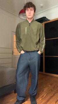 Camicia verde oliva con zip, dal fit rilassato e una tasca frontale, abbinata a jeans in denim blu larghi. Scarpe nere completano l'outfit.