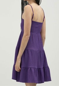 Lila ärmlös klänning med en texturerad bodice, flerskiktad kjol och justerbara axelband. Har en figurnära midja och volangfält.