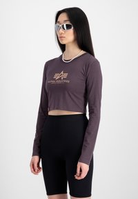 Fialová krátka tričko s dlhým rukávom a zlatým logom, kombinované s čiernymi rebrovanými bicyklovými šortkami. Modelka má na sebe strieborné slnečné okuliare a perlový náhrdelník.