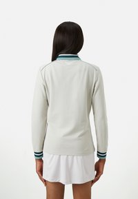 Vrouw met lang donker haar, gekleed in een lichtgrijze langemouwen top en een witte rok, weergegeven van achteren op een eenvoudige witte achtergrond.