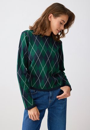 Kobieta w zielonym i granatowym swetrze w romby oraz niebieskich dżinsach, patrząca w dół, z jedną ręką w kieszeni.