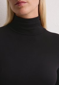 Frau in einem schwarzen gerippten Rollkragenoberteil, das Hals, Untergesicht und oberen Brustbereich vor einer weißen strukturierten Wand zeigt.