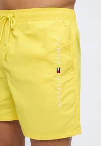 Φωτεινά κίτρινα μαγιό-σορτς με ελαστική μέση και κορδόνι, με κάθετο λογότυπο Tommy Hilfiger και μικρό εμβληματικό σηματάκι στην πλευρά.