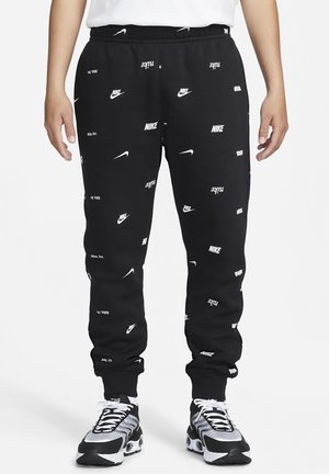 Pantalon de survêtement noir Nike avec logos et virgules blanches Nike dispersés partout, porté avec des chaussures de sport noires et blanches.