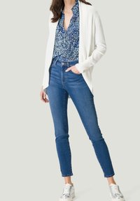 Cardigan blanc sur un chemisier à motifs bleus, associé à un jean ajusté bleu foncé et des baskets argentées. La tenue présente un style décontracté et superposé.