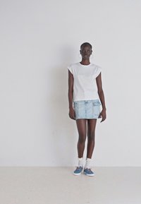 T-Shirt branco de mangas curtas com texto, saia mini em denim azul claro, meias brancas com bordas em renda e sapatilhas azuis. Fundo liso.