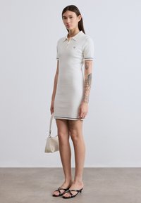 Λευκό φόρεμα πόλο με υφασμένη υφή, κοντά μανίκια και ναυτικούς τόνους. Το μοντέλο κρατάει μια μικρή μπεζ τσάντα, συνδυασμένη με μαύρα σανδάλια.