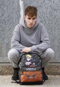 Schwarzer und orangefarbener Rucksack mit einem Grafikdesign von Naruto. Das Material ist Polyester; das Design umfasst Schwerter und einen Charakterdruck auf der Vorderseite.