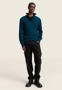 Pulover tricotat de culoare albastru-verzui cu guler cu fermoar, purtat peste o cămașă neagră, asortat cu blugi negri și cizme negre până la gleznă. Textură plată și netedă.