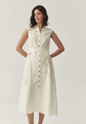 Robe sans manches à boutons en lin crème, ornée de broderies florales sur la gauche et d'une ceinture nouée à la taille. Des boutons décoratifs marron décorent le devant.