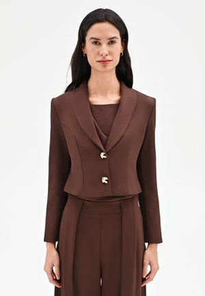 Femme aux longs cheveux foncés portant un blazer marron foncé ajusté avec deux boutons dorés et un pantalon taille haute assorti, debout devant un fond uni.