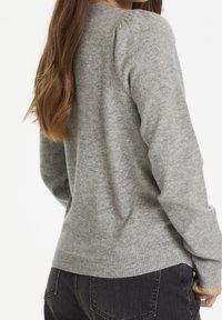 Femme aux longs cheveux bruns portant un pull en maille gris clair et un jean noir, vue de dos sur un fond blanc.
