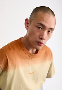 Trumpu rankoviu, apvalios iškirptės marškinėliai, turintys gradientą nuo oranžinės iki grietinėlės, su mažu oranžiniu „Nike“ logotipu ant krūtinės. Pagaminta iš medvilnės.