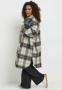 Karierter Button-up-Mantel in Creme, Blau und Braun. Weiche Textur mit großen Knopfverschlüssen und seitlichen Schlitzen. Kombiniert mit dunklen, weiten Hosen.