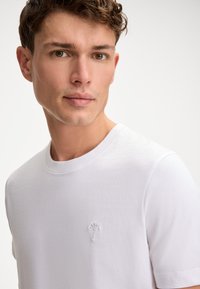 White cotton t-shirt with a classic crew neck, featuring a subtle embroidered logo on the left chest. Smooth texture, short sleeves.