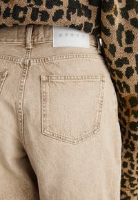 Jeans de mezclilla beige con dos bolsillos traseros, costuras visibles y una etiqueta blanca con caracteres japoneses en la cinturilla.