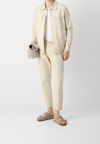 Beżowy zestaw składający się z bawełnianej kurtki i spodni, z białą koszulką polo pod spodem, zestawiony z brązowymi i jasnobeżowymi klapkami. Gładka tekstura i luźny krój.