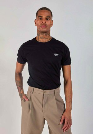 Homme debout contre un fond uni portant un t-shirt noir des Redskins et un pantalon beige, une main dans la poche, avec des tatouages visibles au cou et au bras.
