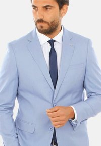 Giacca blu chiaro con un motivo testurizzato, chiusura a un bottone e taschino sul petto, abbinata a una camicia bianca e una cravatta blu navy.