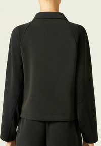 Schwarzer Pullover mit Kragen, langen Raglanärmeln und glatter Textur. Verfügt über einen leicht verkürzten Saum und minimale Nähdetails.