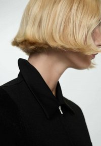 Sort frakke med lynlås og smal krave, vist sammen med en lys blond bob-frisure, der fremhæver glat tekstur og skræddersyet design.