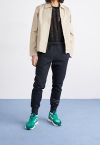 Giacca leggera beige, maglietta nera a maglia strutturata, pantaloni tuta neri con polsini e sneakers verdi con dettagli bianchi e tre strisce.