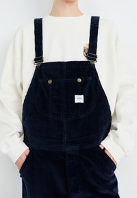Marineblaue Corduroy-Latzhose mit verstellbaren Trägern, Fronttasche und dezentem Lee-Logo-Tag. Kombiniert mit einem hellbeigen Sweatshirt.