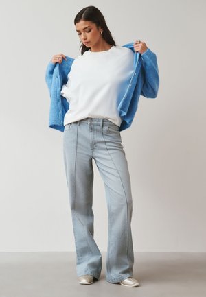 Kobieta w jasnoniebieskich dzwonowatych dżinsach, białej koszulce i niebieskim kardiganie, trzymająca kardigan otwarty na jednolitym jasnym tle.