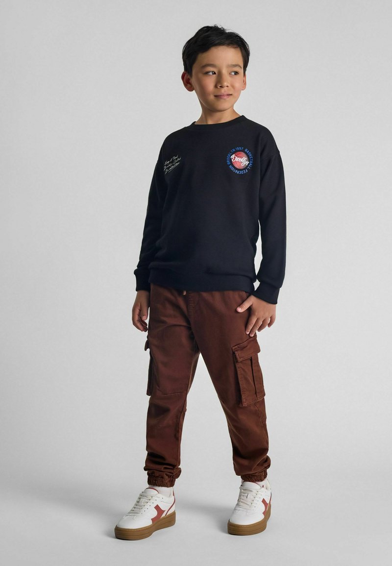 Sudadera negra con logo, pantalones cargo marrones y zapatillas blancas con acentos rojos. Look casual con puños acanalados y bolsillos en los pantalones.