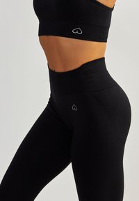 Czarny strój sportowy z dopasowaną górą i legginsami z wysokim stanem. Materiał jest gładki, z subtelnym logo w kształcie serca blisko pasa.