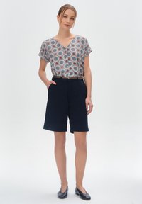 Koszulka z krótkim rękawem w druku z dekoltem w kształcie V i okrągłym wzorem w kolorach czerwonym, niebieskim i białym, w zestawie z granatowymi szortami do kolan i czarnymi płaskimi butami.