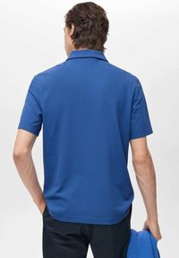 Sininen lyhythihainen polo-paita perinteisellä kauluksella. Silkkinen, yksivärinen kangas; hiukan kaareva helma. Yhdistetty tummansinisiin housuihin ja siniseen neuleeseen.