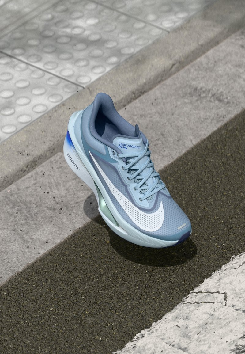 Světlomodrá běžecká bota Nike Zoom Fly s prodyšným síťovým svrškem, výrazným bílým Swooshem a zelenými akcenty na podrážce.