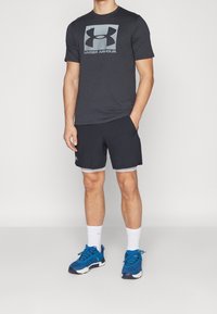 Camiseta de manga corta gris oscuro de Under Armour con logotipo, pantalones cortos negros con ribete gris claro, calcetines blancos y zapatillas deportivas azules.