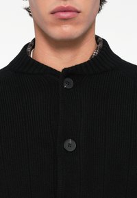 Pogled od blizu na donji dio lica i vrat osobe koja nosi crni rebrasti pleteni kardigan zakopčan na ovratniku preko karirane košulje.