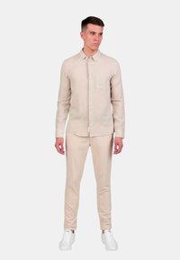 Camicia in lino beige chiaro con colletto button-down, maniche lunghe e taschino sul petto, abbinata a pantaloni su misura dello stesso colore e sneakers bianche.