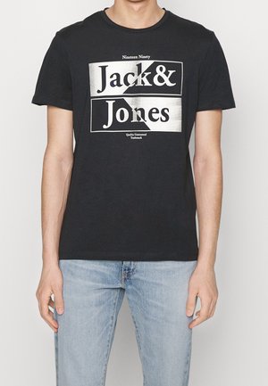 Homme portant un T-shirt noir avec le logo blanc "Jack & Jones" et un jean bleu clair, debout devant un fond uni.