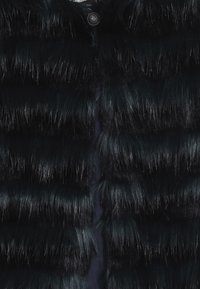 Czarna kamizelka z sztucznego futra o warstwowym designie z długimi, błyszczącymi pasmami. Zapięcie z przodu z okrągłym guzikiem przy szyi.