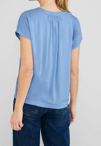 Světle modrá blůza s krátkým rukávem a sklady na zadní straně. Vyrobena z hladké tkaniny, s kulatým výstřihem a volným střihem. V páru s tmavými džíny.