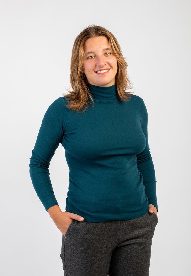 Maglione a collo alto in teal dal design aderente con maniche lunghe, abbinato a pantaloni grigi. Texture liscia senza modelli o decorazioni visibili.